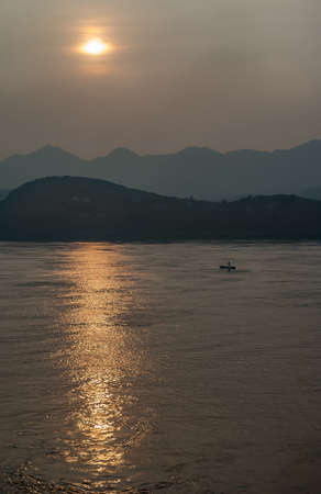 Chongqing, China - May 8, 2010: Evening light on Yangtze River. Small sloop sails on while golden sunset reflected by brown water in front of shoreline with sun above forested hills.の写真素材