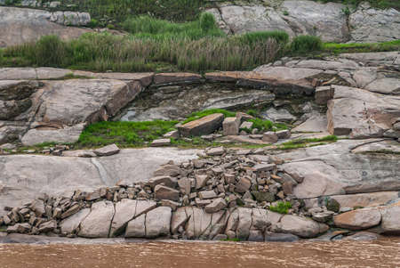 Chongqing, China - May 8, 2010: Evening light on Yangtze River. Closeup of geology of rocky shoreline with patches of green grass and weeds behind brown water.の写真素材