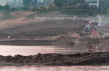 Chongqing, China - May 8, 2010: Evening light on Yangtze River. Site of port construction along green shoreline with houses, and with lots of black dirt, pumping installations, and trucks.の写真素材