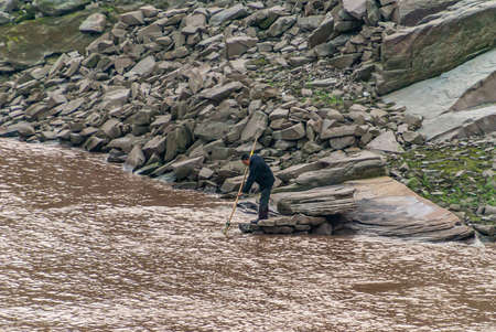 Chongqing, China - May 8, 2010: Evening light on Yangtze River. Man in dark clothes fishes with net on pole from rocky shoreline in brown water. Some green weeds add color.のeditorial素材