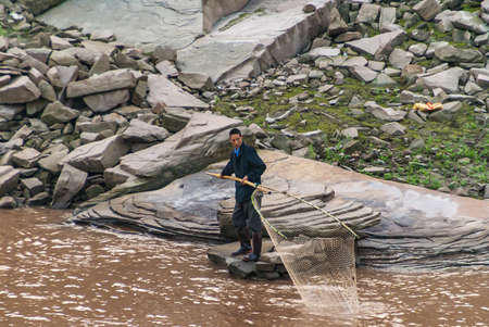 Chongqing, China - May 8, 2010: Evening light on Yangtze River. closeup of man in dark clothes fishing with net on pole from rocky shoreline in brown water. Some green weeds add color.のeditorial素材