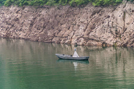 Guandukou, Hubei, China - May 7, 2010: Wu Gorge in Yangtze River: Gray anchored sloop is buoy on green water in front of brown rock shoreline with line of green vegetation.の写真素材