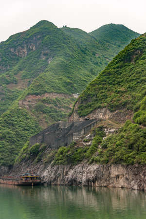 Guandukou, Hubei, China - May 7, 2010: Wu Gorge in Yangtze River. Wall structure above coal loading dock on shoreline of green foliage covered mountain  with barge present on green water.の写真素材