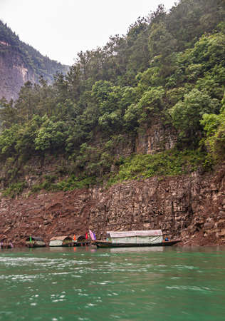 Wushan, Chongqing, China - May 7, 2010: Mini Three Gorges. covered tourist sampans docked against brown cliff rock of green foliage covered mountains behind emerald green water. Flags add color.のeditorial素材