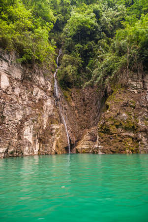 Wushan, Chongqing, China - May 7, 2010: Mini Three Gorges on Daning River. Fine lang white waterfall over brown rock cliff as shoreline behind emerald green water. Foliage on top.のeditorial素材