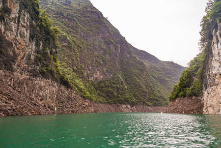 Wushan, Chongqing, China - May 7, 2010: Mini Three Gorges on Daning River. Landscape of wide canyon with high cliff brown rock mountains under silver sky and emerald green water up front.のeditorial素材