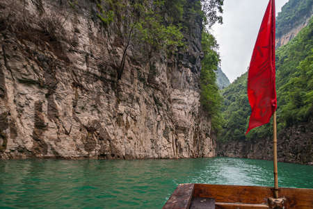 Wushan, Chongqing, China - May 7, 2010: Mini Three Gorges on Daning River. Red flag in emerald green water canyon between brown rock cliffs with some green foliage and patch of silver sky.のeditorial素材
