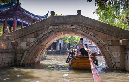 Tongli, JIangsu, China - May 3, 2010: Canal scenery. Captain of brown wooden sculling boat needs to duck his head when sailing under gray stone bridge near blue roof kiosk under brown-greenish water.のeditorial素材