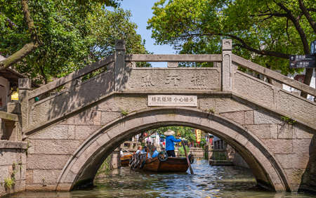 Tongli, JIangsu, China - May 3, 2010: Canal scenery. Closeup of brown-gray stone bow bridge over greenish water, with sculling boat full of tourists on other side. Green foliage and blue sky.のeditorial素材