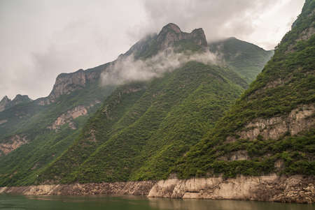 Wushan, Hubei, China - May 7, 2010: Wu Gorge in Yangtze River. Gray cloudscape descends along green forested slopes of mountains into green water. Some brown rocky cliff sections.の写真素材
