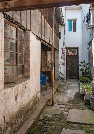 Tongli, JIangsu, China - May 3, 2010: Hutong network of narrow walkways, simple housing, and small businesses. Alley between dirty walls leading to brown door. Plants and blue tricycle.のeditorial素材