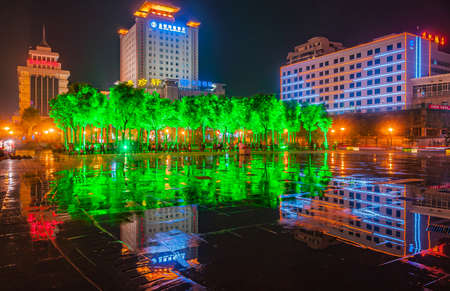 Xian, China - May 1, 2010: Black night landscape. Lighted up HNA Hotel Downtown and other tall buildings seen above park of green lighted trees and reflections in wet surface.のeditorial素材