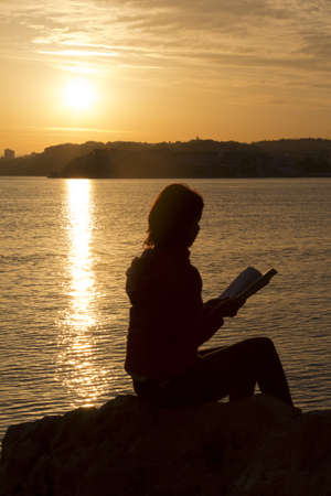 Reading book on the rock near sea at sunset-silhouetteの写真素材