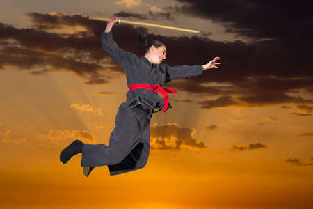 Woman ninja in an aggressive posture flying with katana on sunsetの写真素材