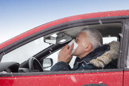 Mid aged man with flu symptoms sneezes in a car on snowy  winter day  Using handkerchief closes mouth with his hands の写真素材