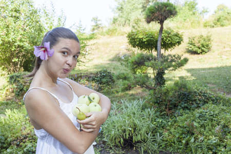Cute natural looking young woman holding organic apples in her beautiful feng shui garden with bonsai pine tree in the background, selective focusの写真素材
