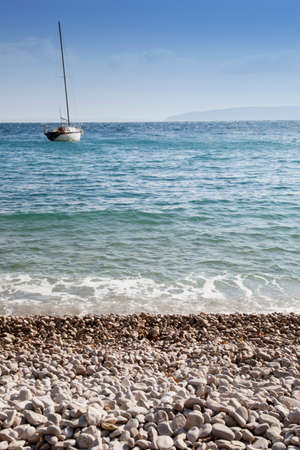 White sandy beach with rough high sea, sail boat and sunny blue sky in the background, selective focus on stones, romantic blue toned, high resolution photoの写真素材