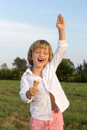 Young cute boy waving hello and eating a tasty ice cream, outdoors on sunny summer afternoonの写真素材