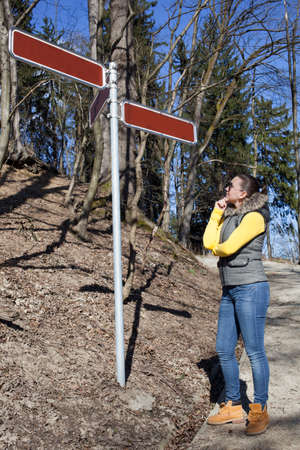 Pretty sporty girl standing at crossroads, under signpost, waypost and trying to decide where to go,...