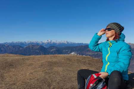 Pretty mid aged woman trekker with backpack resting and sunbathing high in mountains on sunny winter day, against  Mount Triglav, highest mountain in Slovenian  Alps, space for textの写真素材