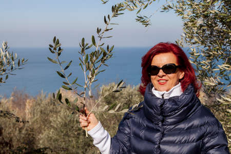 Pretty middle aged woman with red hair meditating, performing joga in beautiful organic olive grove above the sea coast on windy sunny dayの写真素材