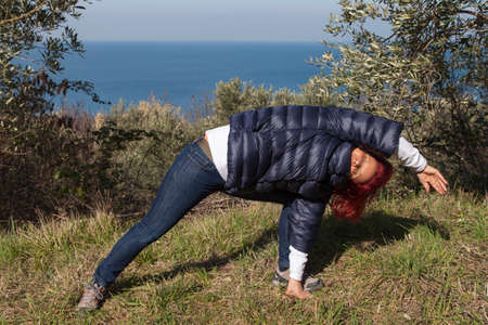 Pretty middle aged woman with red hair meditating, performing joga in beautiful organic olive grove above the sea coast on windy sunny dayの写真素材