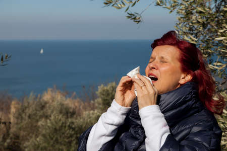 Pretty caucasian middle aged woman with red hair sneezing, blowing nose in handkerchief, outdoors in olive grove above the sea coast, on windy sunny dayの写真素材