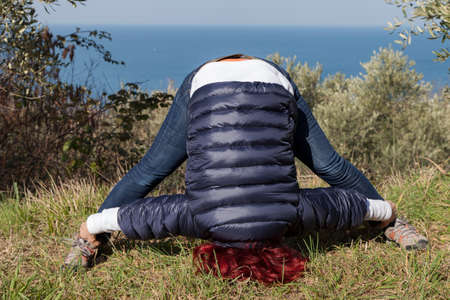 Pretty middle aged woman with red hair meditating, performing joga in beautiful organic olive grove above the sea coast on windy sunny dayの写真素材