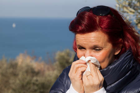 Pretty caucasian woman with red hair sneezing, blowing nose in handkerchief, outdoors in olive grove above the sea coast, on windy sunny dayの写真素材