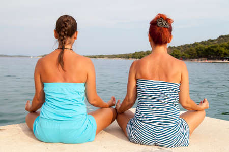 Mother and daughter performing joga on beach, sea coast, meditating in yoga pose on sunny summer dayの写真素材