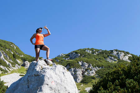 Pretty young woman trekker resting and drinking water, on big rock peak high in the mountains, space for textの写真素材