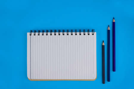 An opened white paper diary beside three wood pencil crayons over a blue paper backgroundの写真素材