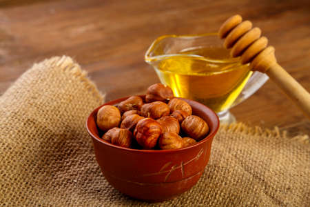 Hazelnuts in a bowl on a sacking next to honey with a spoon on a wooden table.の写真素材