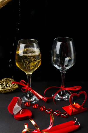 Champagne is poured into glasses tied with a ribbon on a black background, next to a heart, a candlestick with a burning candle and a box with a ring.の写真素材