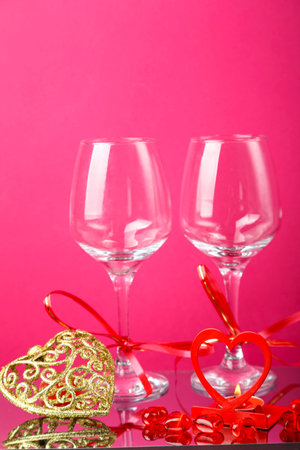 Two champagne glasses with ribbons on a pink background and a gold souvenir heart and a candlestick heart with a burning candle.の写真素材