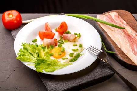 Scrambled eggs with bacon, garnished with tomato and green onion and sliced bacon and tomato and onion on a gray concrete background on a wooden stand.の写真素材
