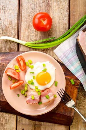 Scrambled eggs with bacon with tomato and onion and on a wooden table in a plate next to chopped bacon and tomatoes.の写真素材