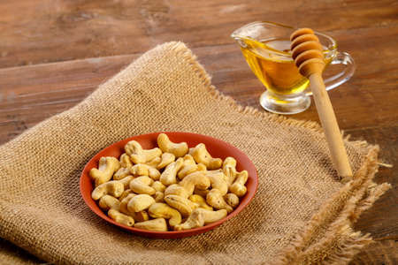 Cashew nuts in a plate on burlap next to honey with a spoon on a wooden table.の写真素材