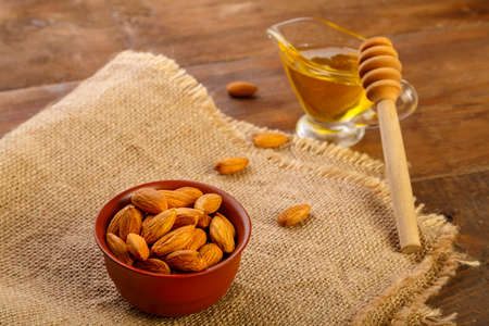 Almonds in a clay cup on sacking next to honey with a spoon on a wooden table.の写真素材