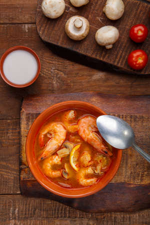 Tom yam soup with shrimps in a plate on a wooden table next to a bowl with coconut milk mushrooms and tomatoes.の写真素材