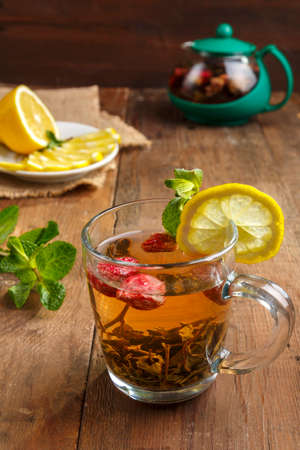 green tea in a glass cup with strawberries mint and lemon on a wooden table and a teapot and lemon in a plate and mint leaves.の写真素材