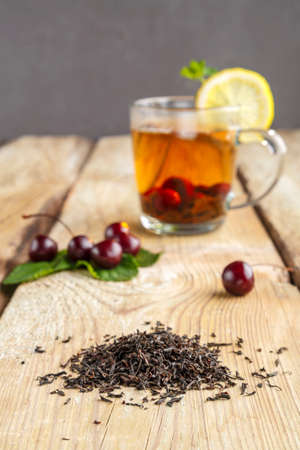 black tea in a glass cup with mint cherries and lemon on a wooden table next to fresh cherries and sprinkled tea leaves.の写真素材