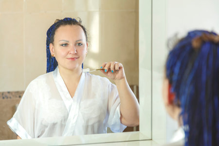 a woman in a white robe with a blue afro hairstyle in the bathroom near the mirror is brushing her teeth.の写真素材