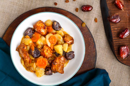 A dish of Jewish cuisine sweet tsimes with carrots, dates and turkey meat in a white plate on a round board on a linen background near dried fruits.の写真素材