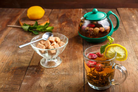 green tea in a glass cup with strawberries mint and lemon on a wooden table and a teapot and brown sugar.の写真素材