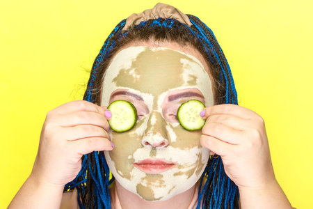 Woman with blue afro braids face in a mask made of green clay with cucumber circles on her eyes on a yellow background.の写真素材