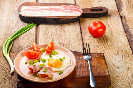 Scrambled eggs with bacon with tomato and onion and on a wooden table in a plate with a fork next to sliced bacon and tomatoes.の写真素材