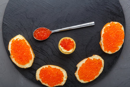 Canapes with butter and red caviar and a spoon with caviar on a black round board on a concrete background copy spaceの写真素材
