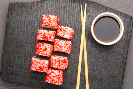 Roll with shrimp and tobiko caviar and sticks on a black board and soy sauce in a sauce bowl on a gray background.の写真素材