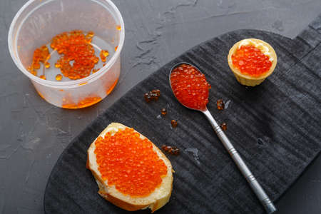 Tartlet with butter and red caviar on a black round board next to a spoon with caviar.の写真素材